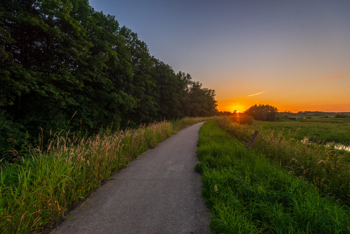 Een verhard en verhoogd wandelpad dat doorsnijdt in het landschap tijdens een kleurrijke zonsondergang